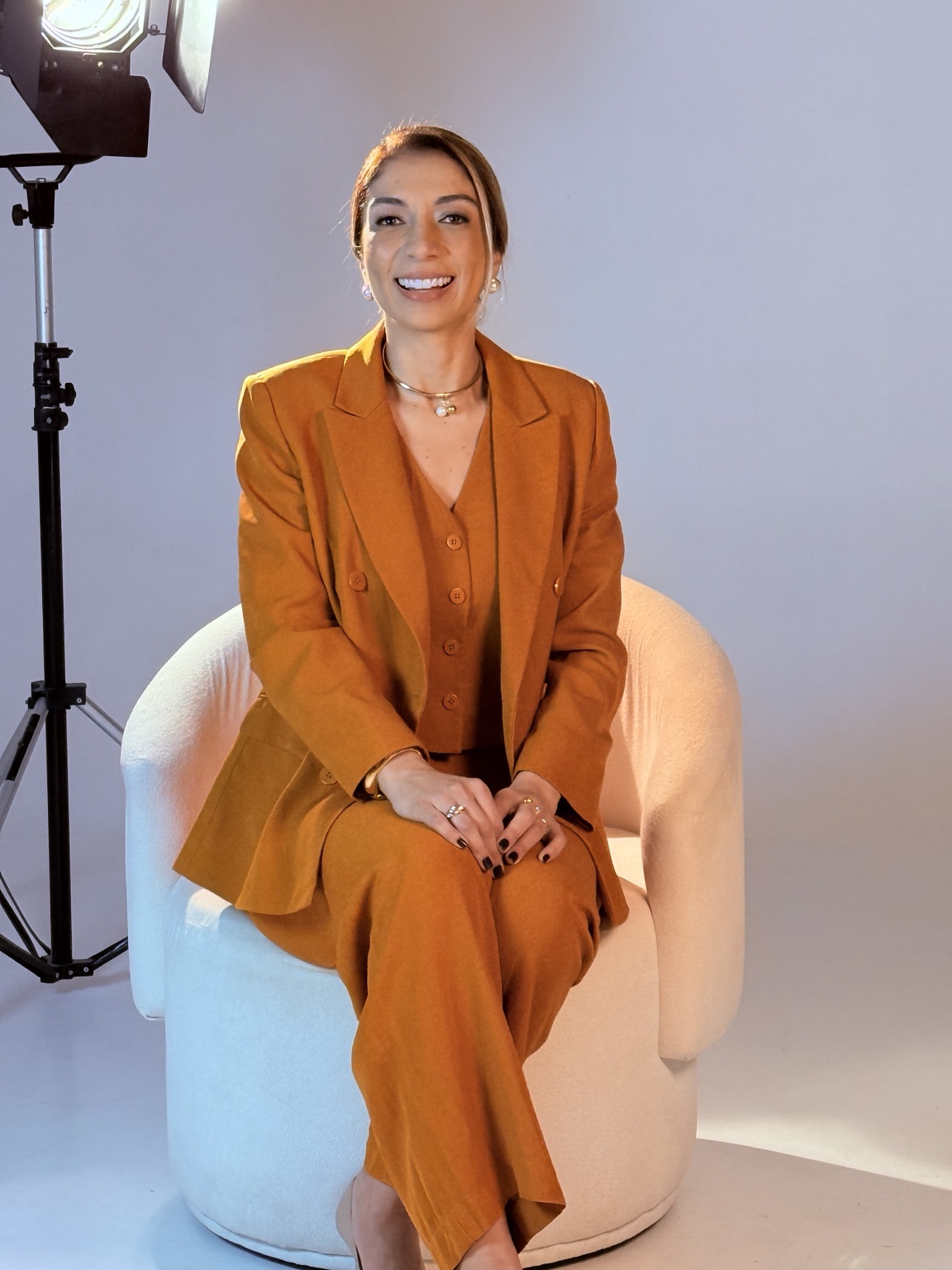 Carla Modesto sorrindo, sentada em uma poltrona clara.